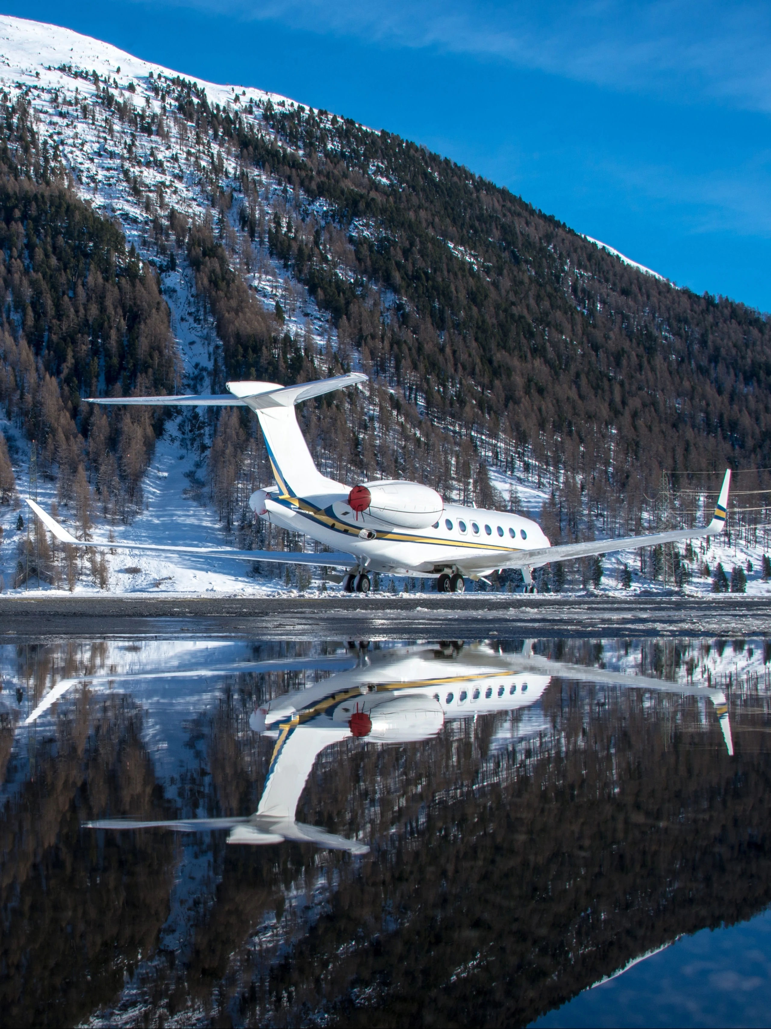 A private jet is parked on a snowy runway, with a mountain backdrop and its reflection visible in a nearby puddle. The scene captures the contrast between the sleek aircraft and the rugged, snow-covered landscape under a clear blue sky.